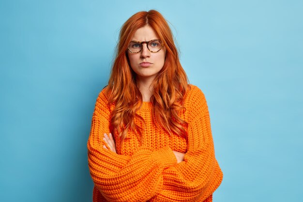 Beledigde boze jonge vrouw voelt zich ongelukkig en geïrriteerd houdt armen over elkaar, is ontevreden over iets, houdt armen over elkaar en wacht op uitleg, draagt oranje wintertrui. Negatieve gevoelens