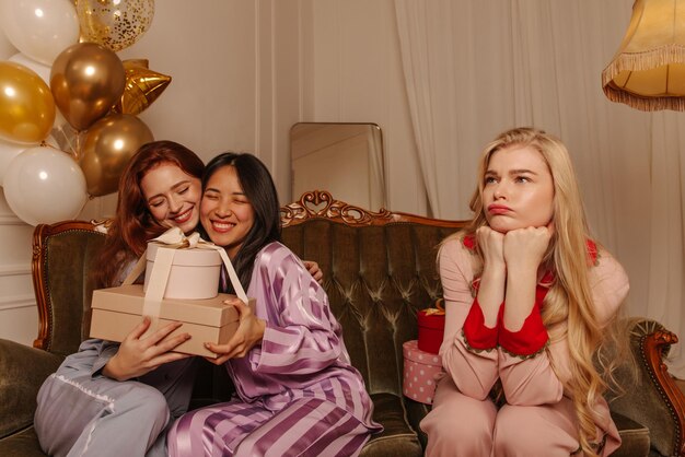 Beledigde jonge blonde vrouw in pyjama zit op de bank naast vrolijke vriendinnen met geschenkdozen Vrije tijd en lifestyle concept