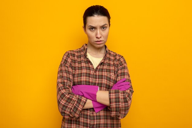 Beledigde jonge schoonmaakster in vrijetijdskleding in rubberen handschoenen met een fronsend gezicht met gekruiste armen over oranje muur