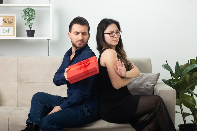 Beledigde knappe man die geschenkdoos geeft aan ontevreden mooie jonge vrouw in optische bril zittend op de bank in de woonkamer op maart internationale vrouwendag