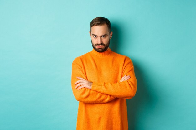 Beledigde man kijkt boos naar je, armen gekruist op de borst en boos staren, staande in oranje trui tegen turkooizen muur.