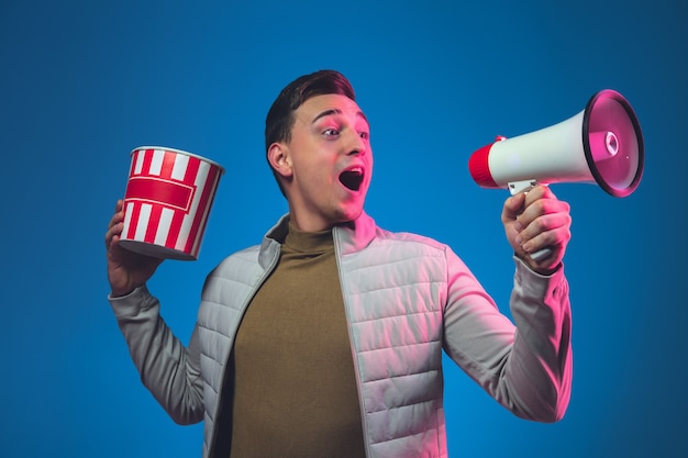 Bellen met speaker en popcorn. portret van een blanke man geïsoleerd op blauwe muur in roze neonlicht.