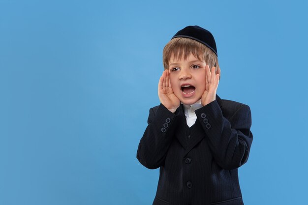 Bellen, schreeuwen. Portret van een jonge orthodoxe joodse jongen geïsoleerd op blauwe muur. Purim, zaken, festival, vakantie, jeugd, viering Pesach of Pesach, jodendom, religieconcept.