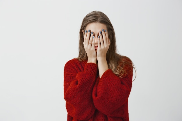 Beloof het, ik gluur niet. schot van vermoeide en beu aantrekkelijke vrouw in rode losse trui, bedekkend gezicht met handpalmen, gestrest en uitgeput gevoel, moet ontspannen en slapen over grijze muur