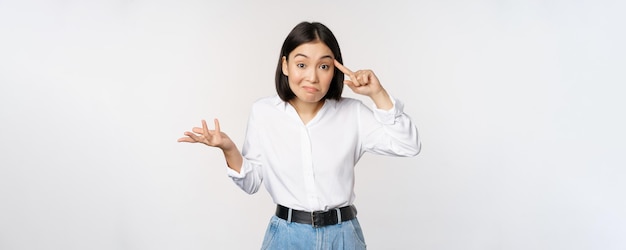 Ben je krankzinnig Portret van gefrustreerde jonge aziatische vrouw die met haar vinger in de buurt van het hoofd rolt en iemand gek of dom op een witte achtergrond staat