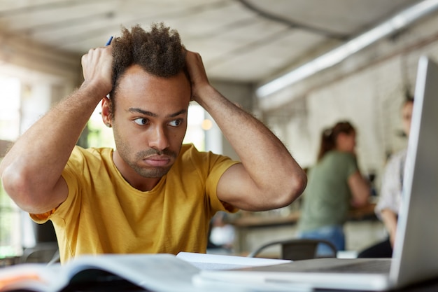 Gratis foto benadrukt jonge afro-amerikaanse student gefrustreerd gevoel, zittend op coworking space voor open laptop computer, hoofd met handen vast te houden