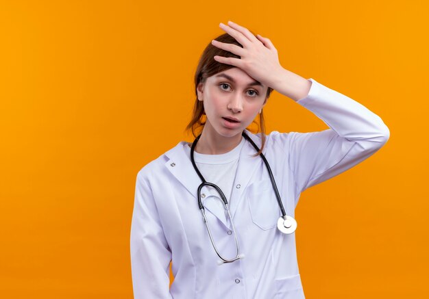 Benadrukt jonge vrouwelijke arts die medische mantel en stethoscoop draagt die hand op hoofd op geïsoleerde oranje muur met exemplaarruimte zet