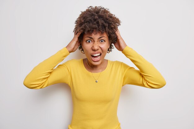 Beschaamd krullend haired vrouw grijpt hoofd roept luid voelt geïrriteerd weet niet hoe moeilijke situatie op te lossen draagt gele trui geïsoleerd op witte achtergrond Negatieve emoties