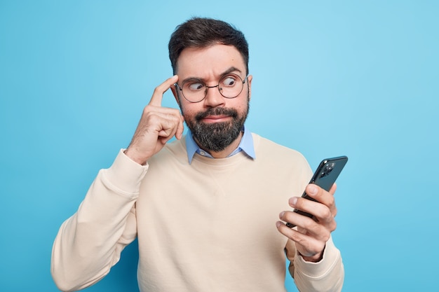 Beschaamde bebaarde man gericht op smartphone houdt vinger op tempel probeert zich te concentreren op informatie staart naar display gekleed in nette trui poses tegen blauwe muur. Technologie concept