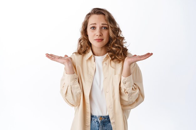 Besluiteloos en verward blond meisje, schouders ophalend met terughoudend gezicht, weet niet wat doen, verbaasd tegen de witte muur staan