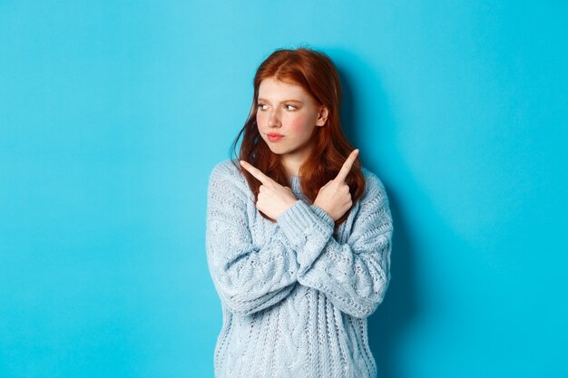 Besluiteloos roodharige tienermeisje dat een beslissing neemt, de vingers zijwaarts wijst en twijfelachtig naar links kijkt, staande in een trui tegen een blauwe achtergrond