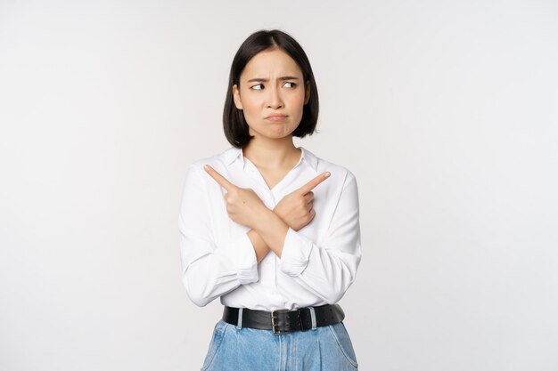 Besluiteloze aziatische vrouw die met de vingers zijwaarts wijst, met de vingers wijst en er geen idee van heeft, verward met keuzes die op een witte achtergrond staan