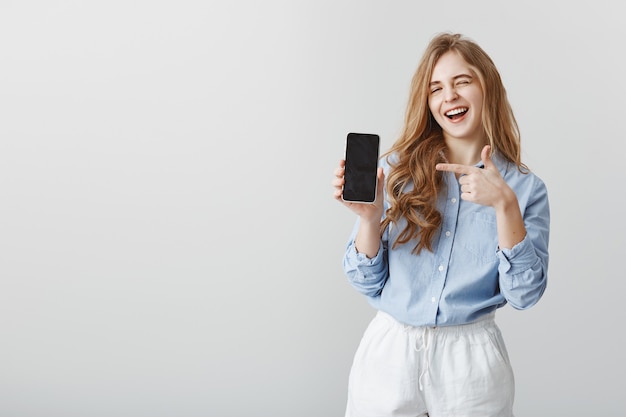 Beste keuze ooit. Portret van knap Kaukasisch vrouwelijk model met blond haar in blauwe blouse, knipogen en glimlachen terwijl smartphone wordt weergegeven en met wijsvinger naar apparaat wijst, bevordering van