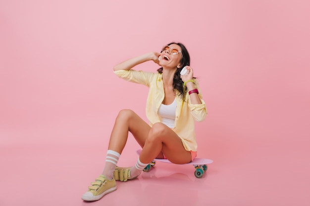 Betoverend donkerbruin meisje met gebruinde huid zittend op een longboard met gekruiste benen. Indoor portret van romantische Spaanse vrouw in gele sneakers luisteren muziek in koptelefoon.