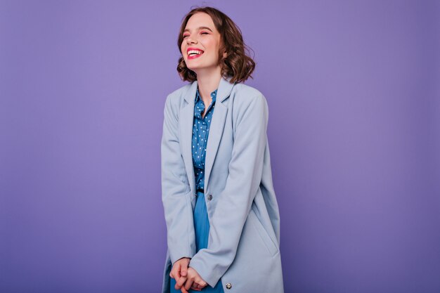 Betoverend krullend meisje dat met gesloten ogen op purpere muur lacht. Indoor foto van mooie brunette dame draagt elegante blauwe jas.