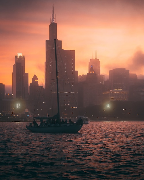 Gratis foto betoverend uitzicht op de boot in de oceaan en de silhouetten van hoge gebouwen tijdens zonsondergang