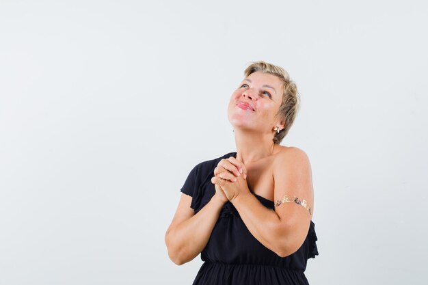 Betoverende vrouw die gecombineerde handen op haar borst houdt terwijl zij in zwarte blouse wenst en hoopvol kijkt. vooraanzicht.