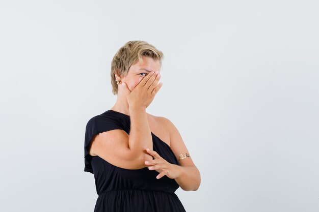 Betoverende vrouw die hand op gezicht houdt terwijl over haar hand in zwarte blouse kijkt en bang kijkt. vooraanzicht.