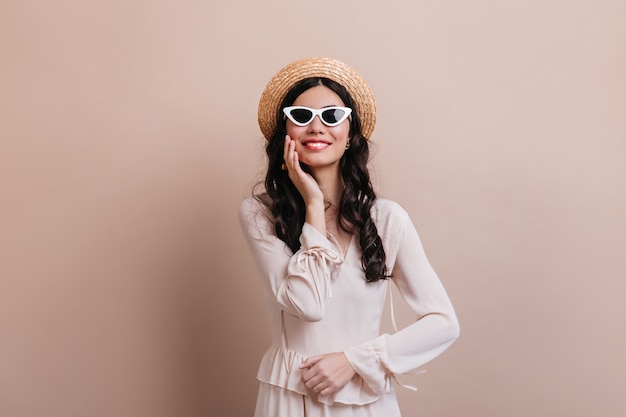 Betoverende vrouw die in zonnebril op beige achtergrond lacht. Vooraanzicht van tevreden donkerbruine vrouw in strohoed.