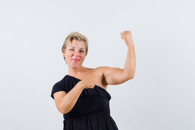 Betoverende vrouw die naar haar armspieren in zwarte blouse richt en tevreden kijkt. vooraanzicht.