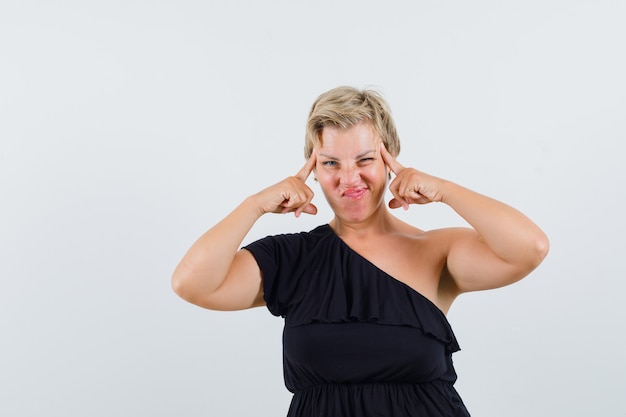 Betoverende vrouw die op haar tempels in zwarte blouse richt en er slim uitziet. vooraanzicht.