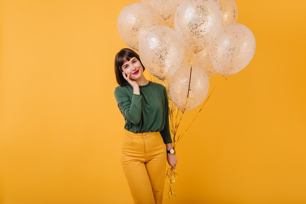 Betoverende vrouw met steil haar poseren met een heleboel sprankelende ballonnen. Binnen schot van glimlachend zorgeloos meisje in groene trui en gele broek.