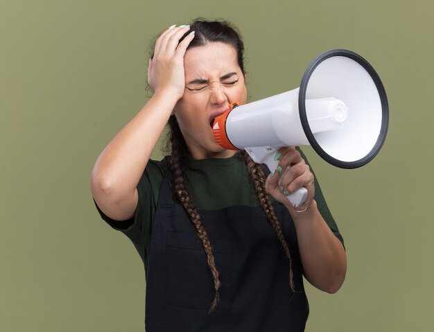 Betreurd met gesloten ogen jonge vrouwelijke kapper in uniform spreekt op luidspreker hand op hoofd zetten geïsoleerd op olijfgroene muur