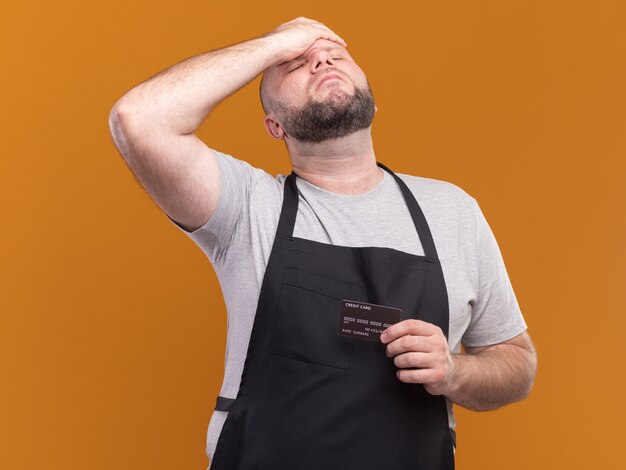 Betreurd met gesloten ogen Slavische mannelijke kapper van middelbare leeftijd in uniform met creditcard en hand op voorhoofd geïsoleerd op oranje muur