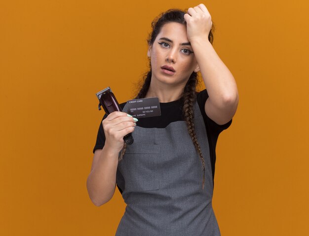 Betreurde jonge vrouwelijke kapper in uniform bedrijf creditcard en tondeuse hand zetten voorhoofd geïsoleerd op oranje muur met kopie ruimte