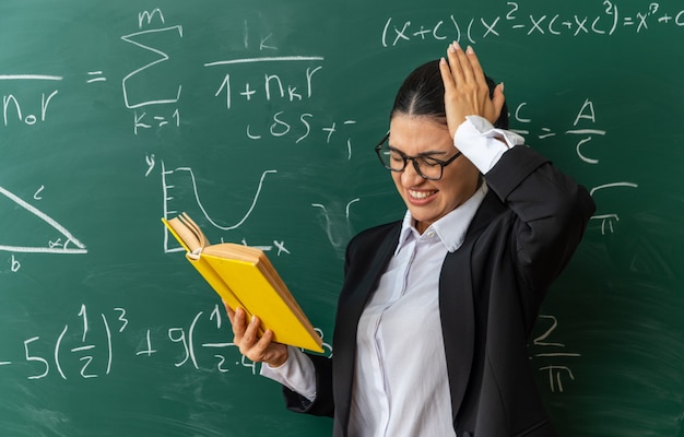 Betreurde jonge vrouwelijke leraar die een bril draagt die vooraan het leesboek van het schoolbord staat en de hand op het hoofd zet in de klas