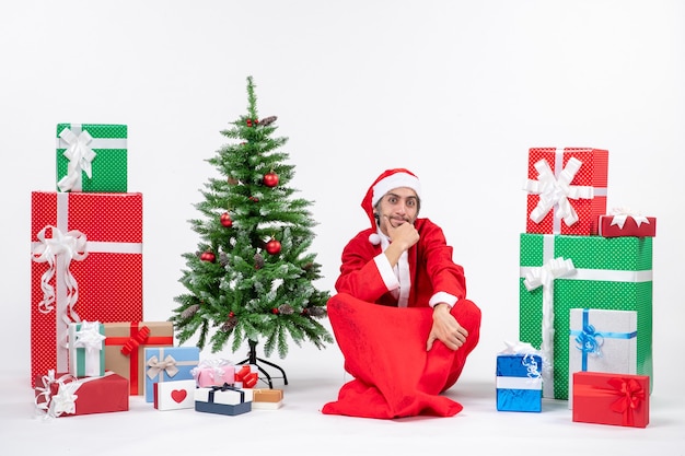 Betrokken doordachte jonge volwassene verkleed als kerstman met geschenken en versierde kerstboom zittend op de grond op witte achtergrond