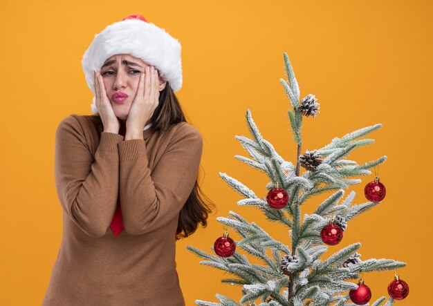 Betrokken jong mooi meisje met kerstmuts met stropdas staande in de buurt van kerstboom handen op wangen geïsoleerd op oranje muur zetten