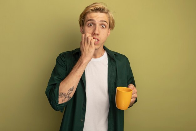 Betrokken jonge blonde kerel die groene t-shirt draagt die kop thee houdt en nagels bijt