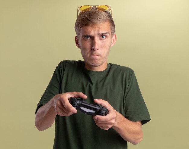 Betrokken jonge knappe kerel die een groen shirt met een bril op het hoofd draagt en op de joystick van de gamecontroller speelt die op olijfgroene muur wordt geïsoleerd