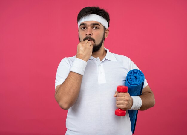 Betrokken jonge sportieve man met hoofdband en polsband halter met yoga mat hand op kin te houden