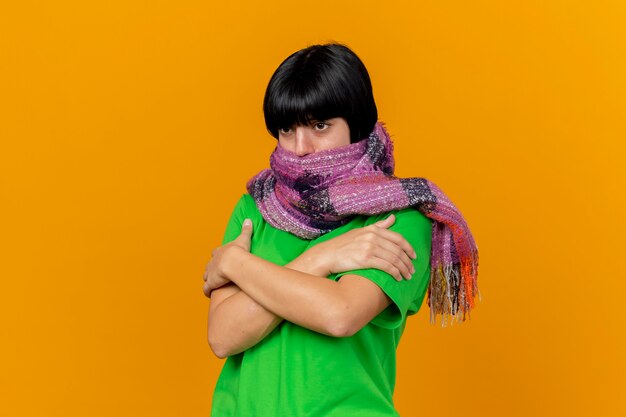 Bevriezende koude jonge zieke vrouw die sjaal draagt die mond bedekt met sjaal die handen op wapens houdt die op oranje muur worden gekruist die recht kijken