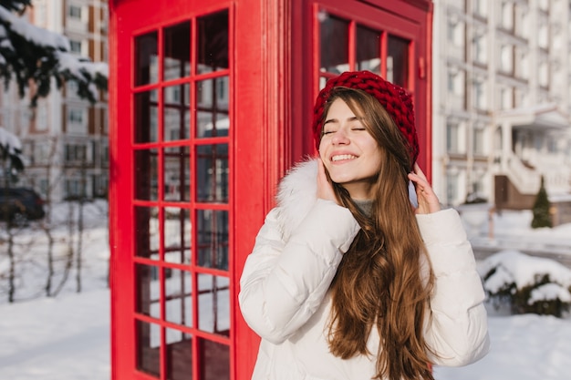 Bevroren zonnige sneeuw ochtend van vrolijke jonge vrouw met lang donkerbruin haar in rode hoed genieten van wintertijd in de buurt van rode telefooncel op straat. Chillen in de zon, glimlachend met gesloten ogen.