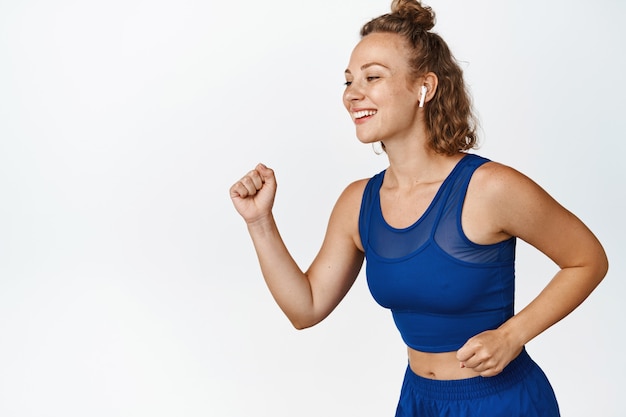 Bewegingsportret van fitnessvrouw in koptelefoon die rent, jogt met een lachend blij gezicht, sportkleding draagt op wit