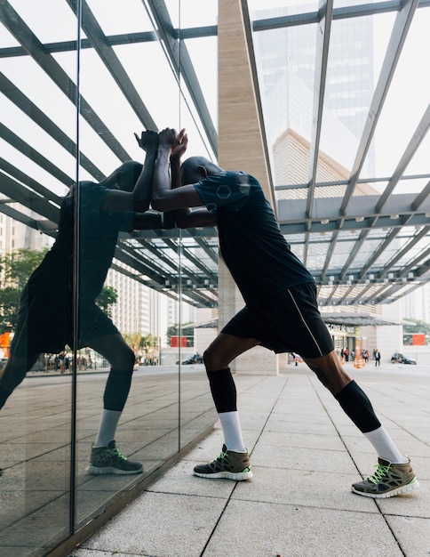 Gratis foto bezinning van mannelijke atleet die zijn spier uitrekt die op weerspiegelend glas leunt