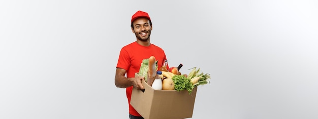 Bezorgconcept Knappe Afro-Amerikaanse bezorger met pakketdoos met kruidenierswaren en drinken uit de winkel Geïsoleerd op grijze studioachtergrond Kopieer ruimte