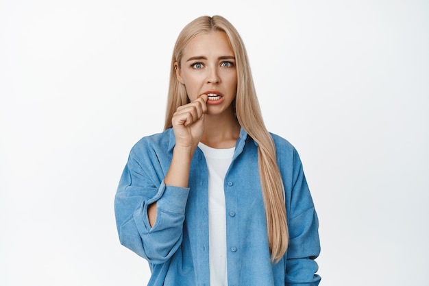 Bezorgd blond meisje bijt in haar vinger en kijkt schuldig naar de camera die angstig staat tegen een witte achtergrond