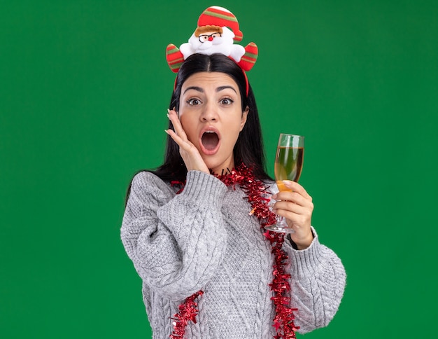 Bezorgd jong kaukasisch meisje met de hoofdband van de kerstman en klatergoudslinger om de nek met een glas champagne met de hand op het gezicht geïsoleerd op een groene muur met kopieerruimte