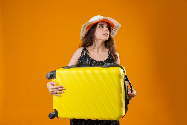 Bezorgd jong mooi reizigersmeisje in kleding in polka dot in de holdingskoffer van de de zomerhoed die opzij status over gele achtergrond kijken