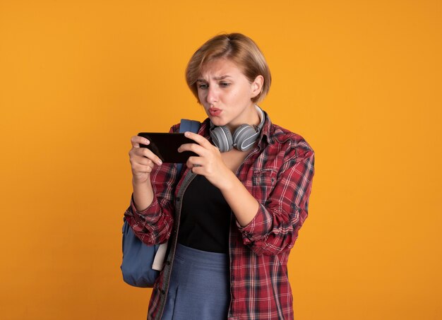 Bezorgd jong slavisch studentenmeisje met hoofdtelefoons die rugzak dragen die houden en naar telefoon kijken