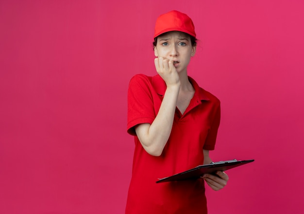 Bezorgd jonge mooie levering meisje in rood uniform en pet houden Klembord kijken camera en bijten vingers geïsoleerd op karmozijnrode achtergrond met kopie ruimte