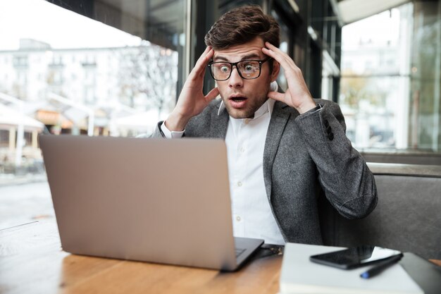 Bezorgd verwarde zakenman in brillen zitten door de tafel in café terwijl hoofd en laptop computer kijken