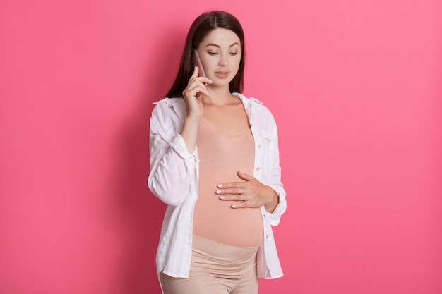 Bezorgd zwangere vrouw praten aan de telefoon met haar arts, wordt geconcentreerd, houdt de hand op haar buik, permanent geïsoleerd op roze achtergrond, jurken casual kleding.