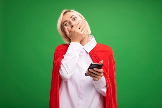 Bezorgde blonde superheldenvrouw van middelbare leeftijd in rode cape met mobiele telefoon die hand op mond houdt geïsoleerd op groene muur met kopieerruimte