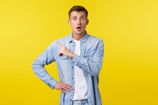 Bezorgde en bezorgde blonde knappe man die er onzeker en angstig uitziet, wijzende vinger in de linkerbovenhoek staren camera gealarmeerd, bang als hij over een gele achtergrond staat.