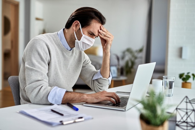 Bezorgde freelance werknemer die beschermend gezichtsmasker draagt tijdens het lezen van problematische e-mail op een computer en thuis werken tijdens virusepidemie